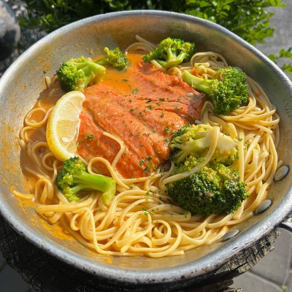 Salmon and broccoli pasta in a metal pan with a lemon wedge, against a green background.