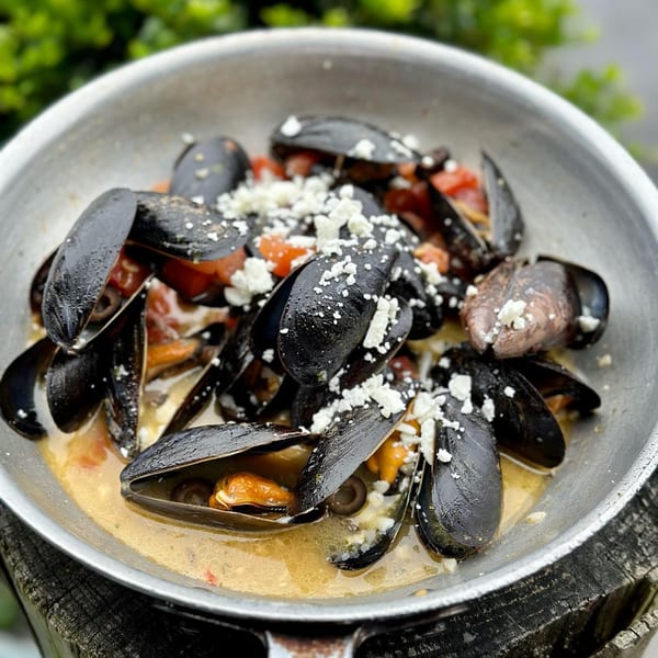 Steamed mussels in a pan with broth, diced tomatoes, and crumbled feta cheese.