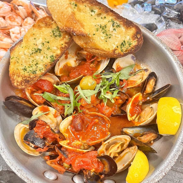 Steaming bowl of Shore Fresh clams and mussels in red sauce, garnished with basil, lemon, and garlic bread.